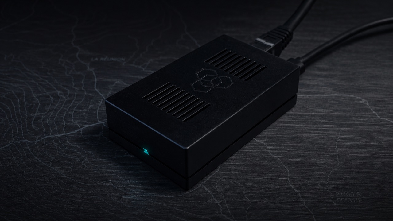 Sonde RIPE Atlas, boîtier USB noir avec LED teal, posée sur une carte topographique sombre.